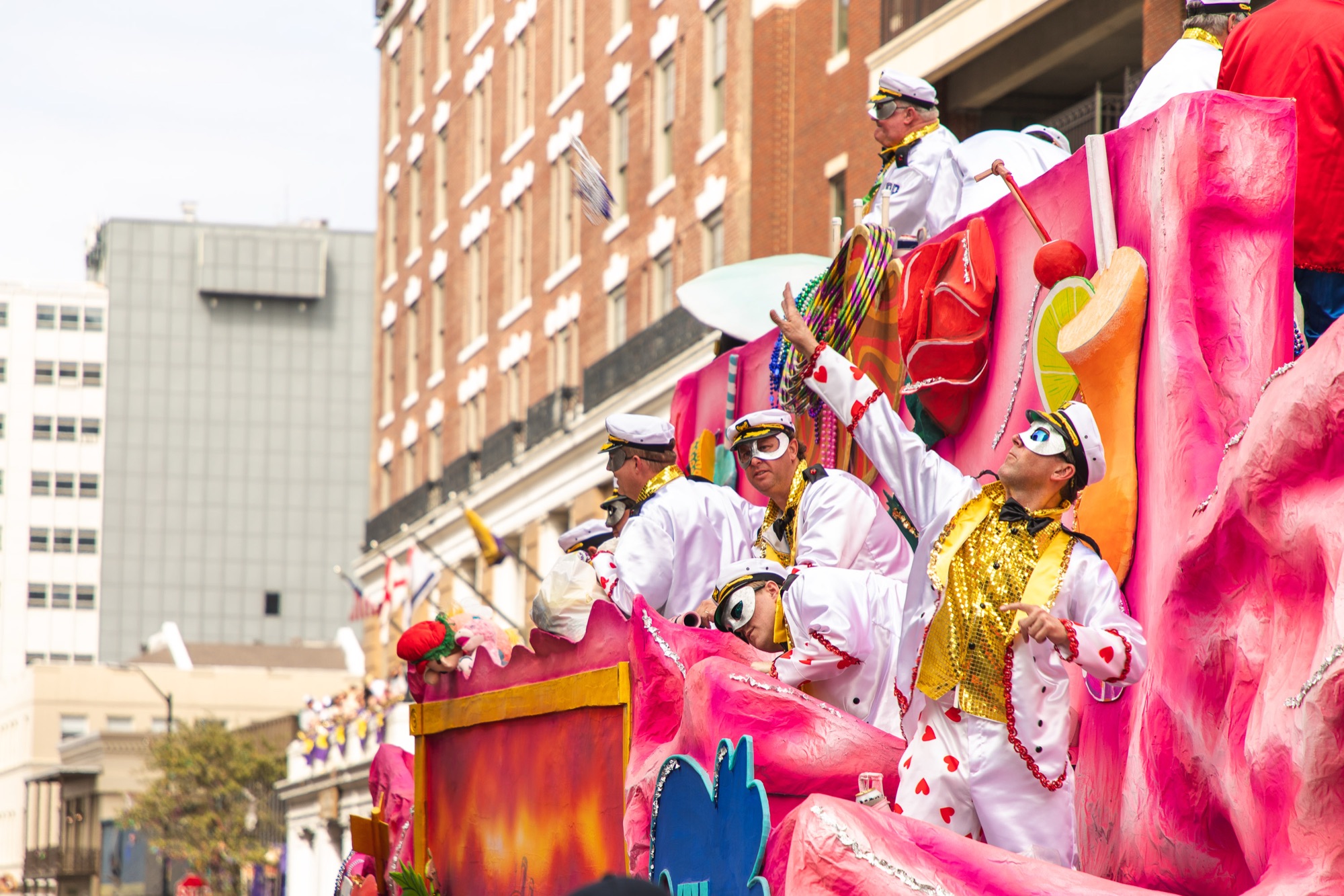 Masked riders in sailor caps throw beads from a pink float
