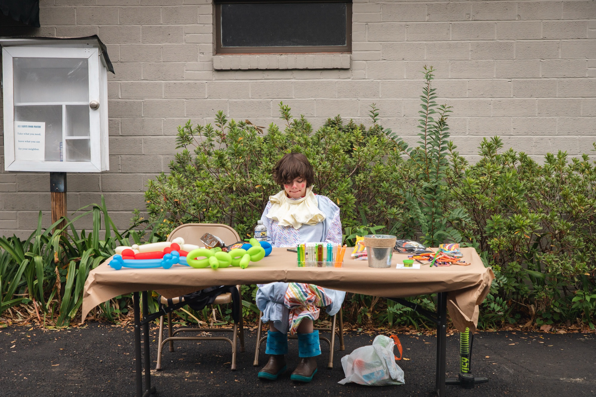 A kid in a clown costume sits alone at a balloon-animal table
